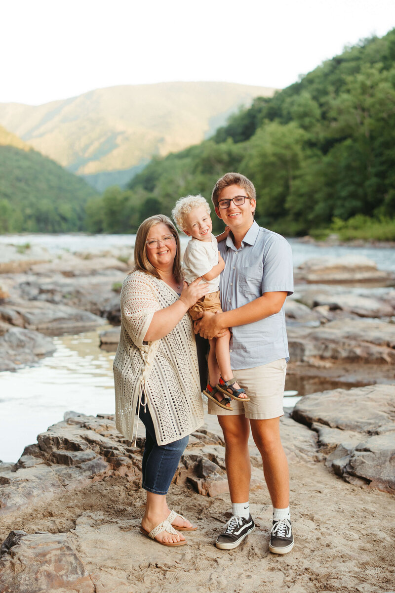 April Booher hugs her toddler son in a plaid shirt and jeans on the edge of a lake in fall at sunset.