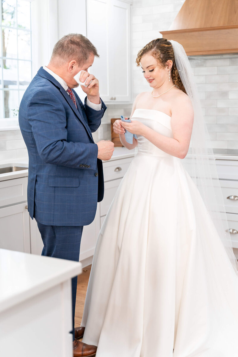 A groom in a blue suit wipes away tears while standing in a kitchen with his bride.