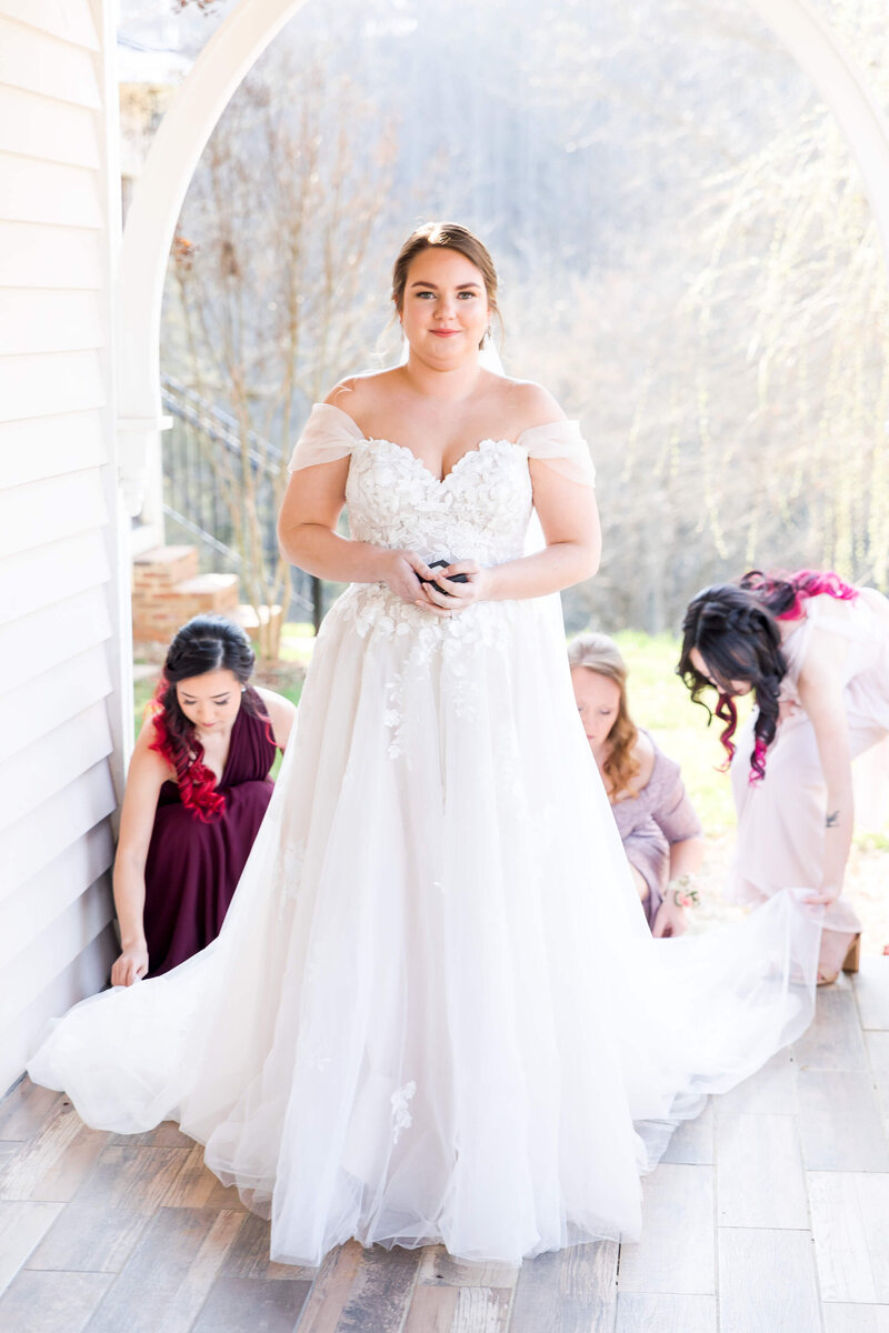 A bride stands smiling on a porch as her bridesmaids adjust her train behind her for a Johnson City wedding photographer.