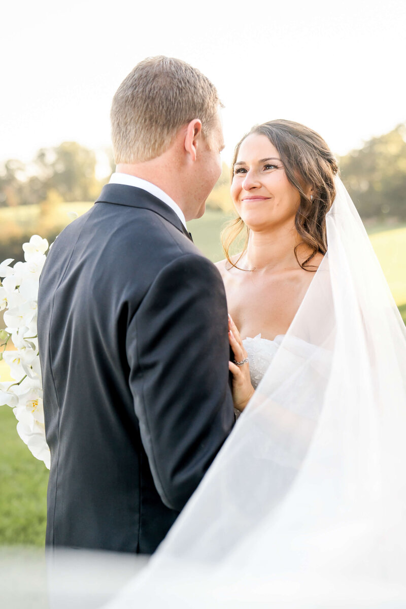 A happy bride smiles at her groom in a black tux while out in the sunset with veil flying around them.