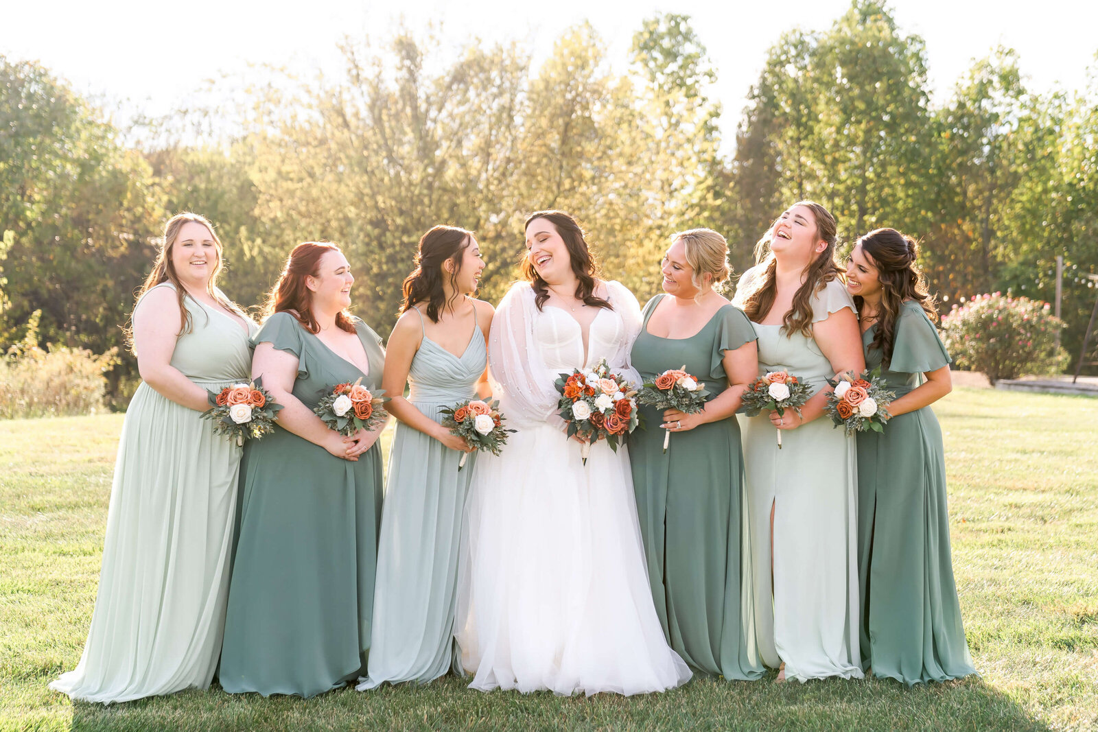 A happy bride laughs while standing with her bridesmaids in sage green in a field at sunset" title="Bride Laughing With Bridesmaids.