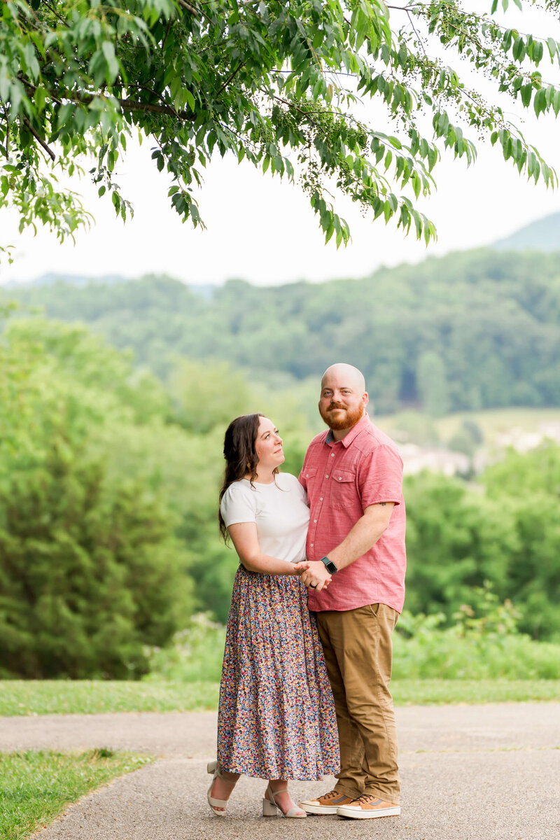 A happy engaged couple hold hands and smile under a tree on a sidewalk.