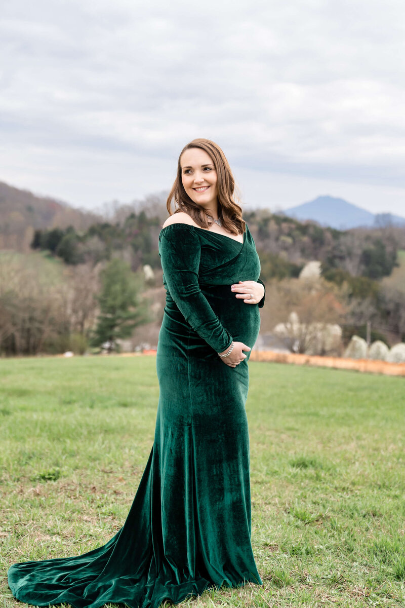 An expecting couple in green and brown snuggle holding hands under the bump in a park in the fall captured by their Johnson City maternity photographer.