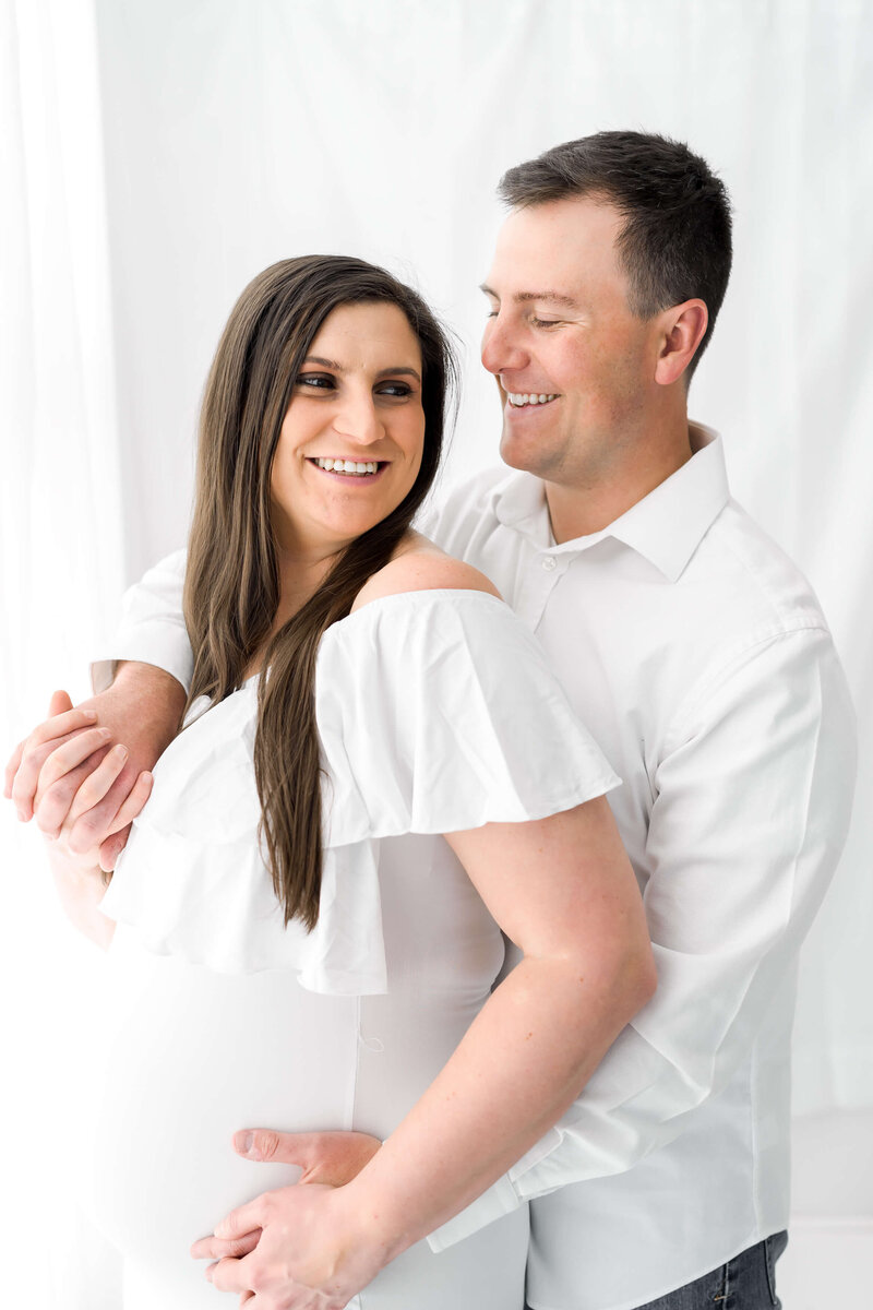 A couple kiss while holding hands on the bump both in white outfits in a studio.