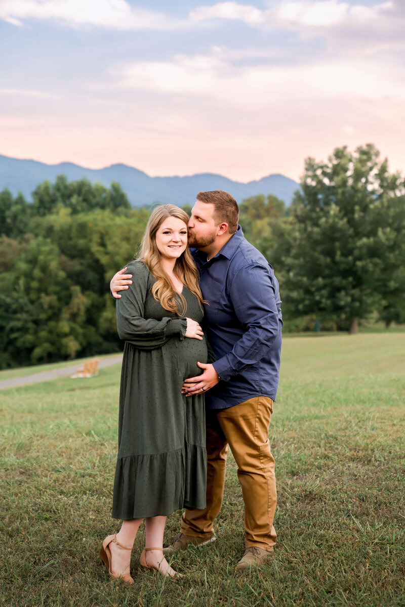 A happy pregnant couple stand on a meadow in the mountains as the man kisses her cheek.