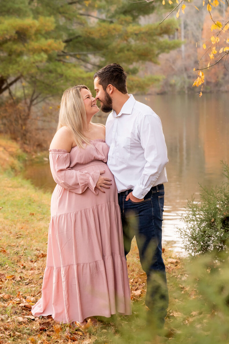 A happy pregnant couple lean in for a kiss by the water captured by a Johnson City maternity photographer.