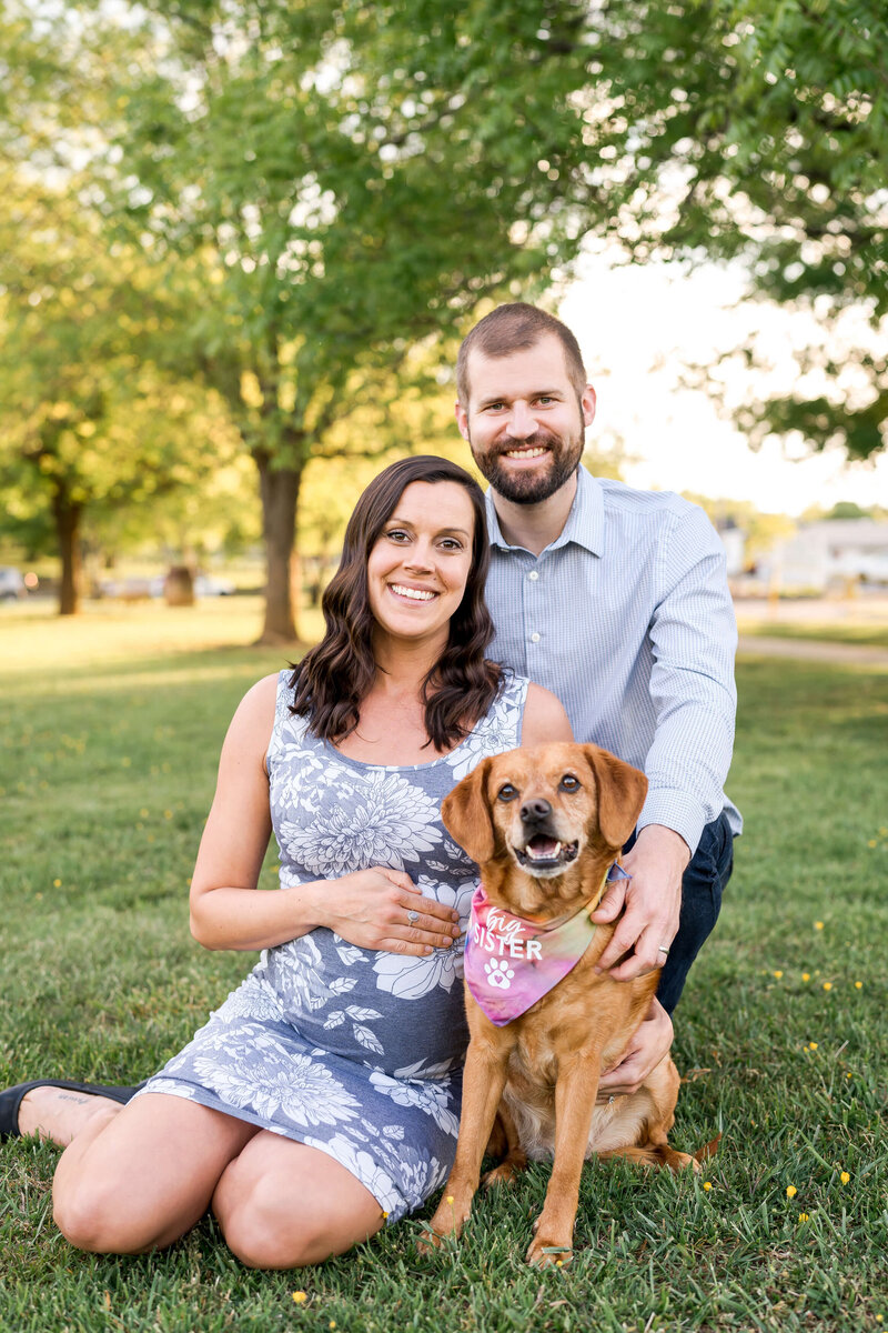 An expecting couple sit in a park lawn with their small brown dog wearing a pink bandana.