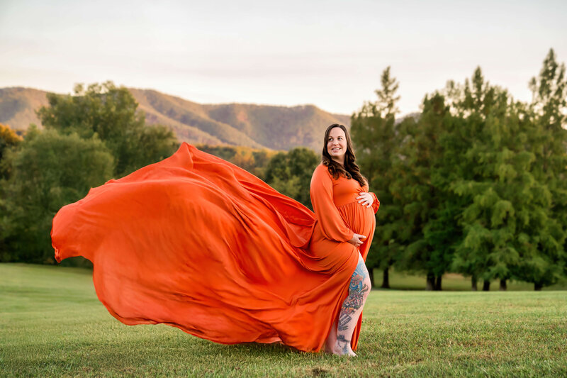 A mom to be stands in a park lawn with the train of her orange maternity gown blowing in the wind behind her captured by a Johnson City maternity photographer.