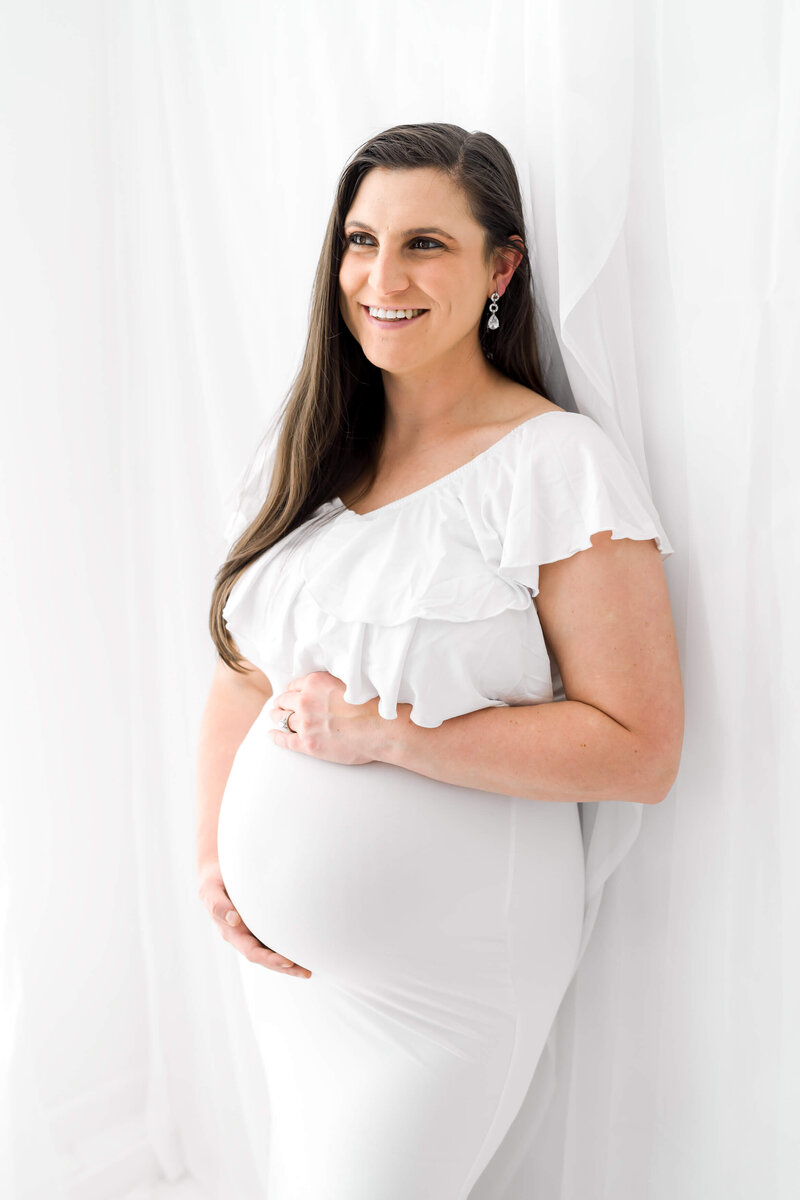 A smiling pregnant woman leans against a white wall in a white maternity gown.