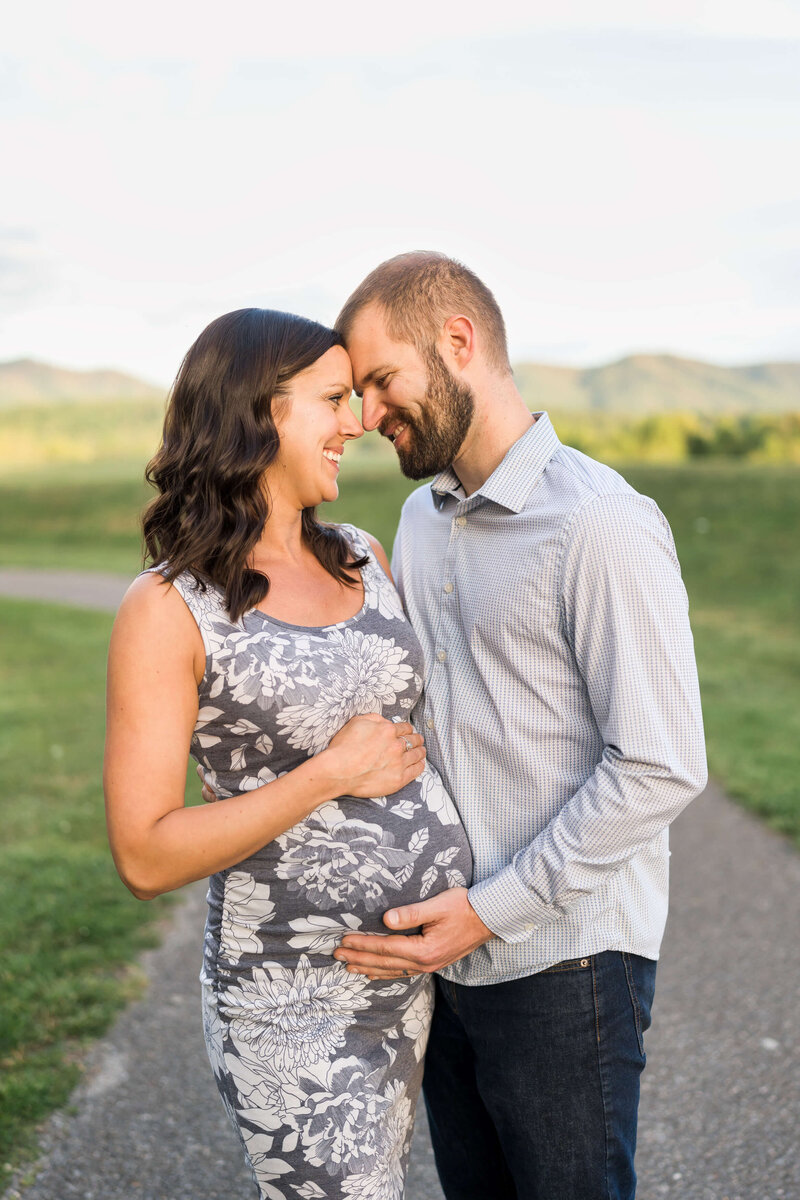A mom to be in a grey flower print maternity dress touches foreheads and holds the bump with her husband in grey in a park sidewalk.