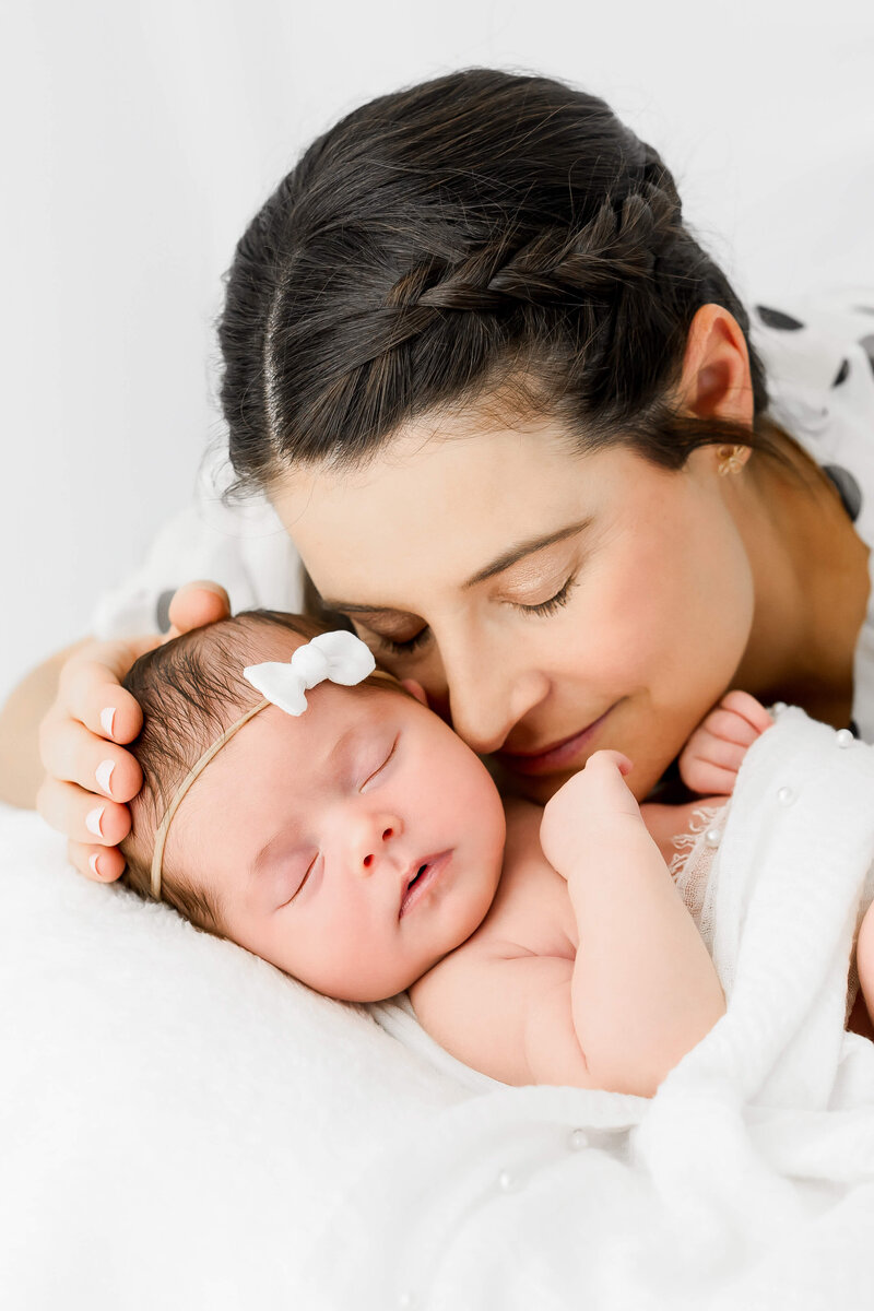 A new mom with dark hair snuggles her sleeping newborn cheek to cheek on a white bed.
