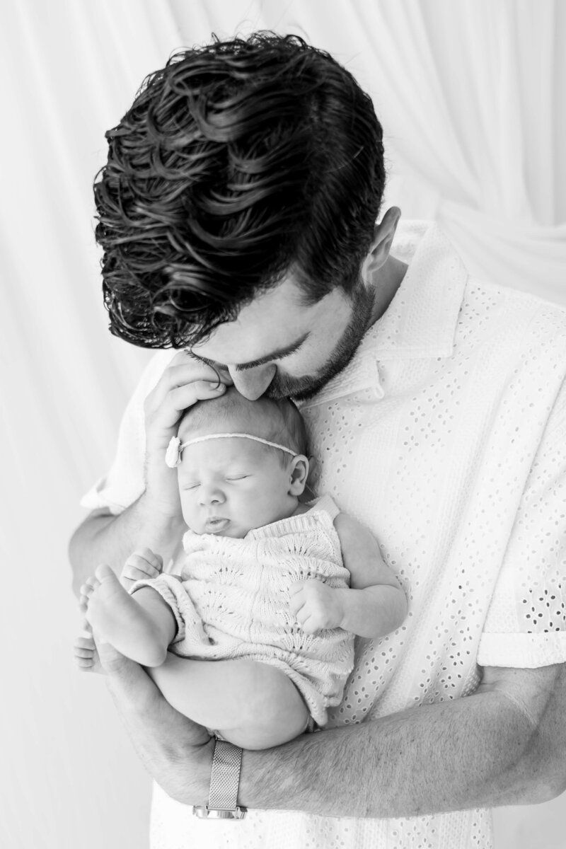 A new dad in a white shirt snuggles his sleeping newborn daughter against his chest in black and white during a Johnson City newborn photography session.