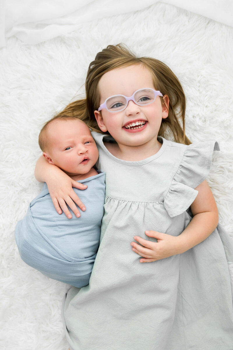 A toddler girl in a dress lays on a white bed snuggling her newborn baby brother in a Johnson City newborn photographer's studio.