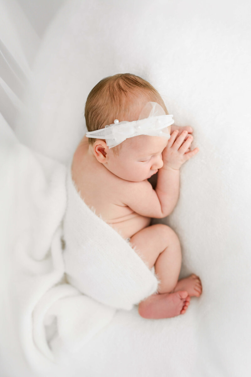 A newborn baby girl with a white headband sleeps on a white bed in the studio of a Johnson City newborn photographer.