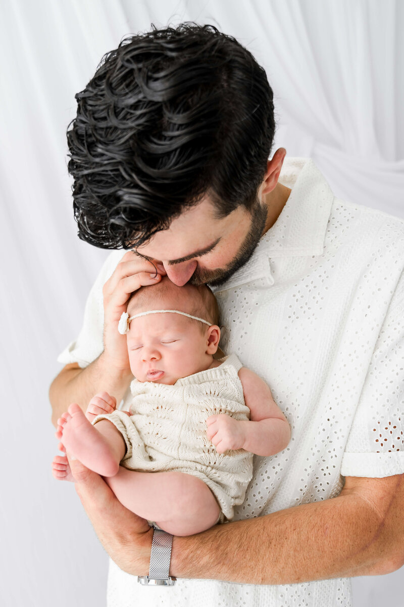 A new dad in a white polo cradles her sleeping newborn against his chest in a white onesie.