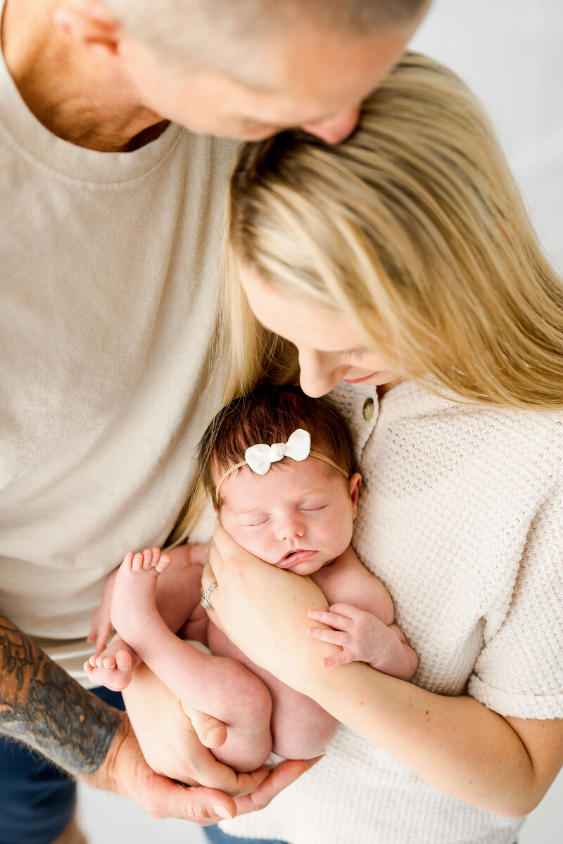 A newborn with a white bow sleeps against mom's chest in dad's hands as they snuggle.