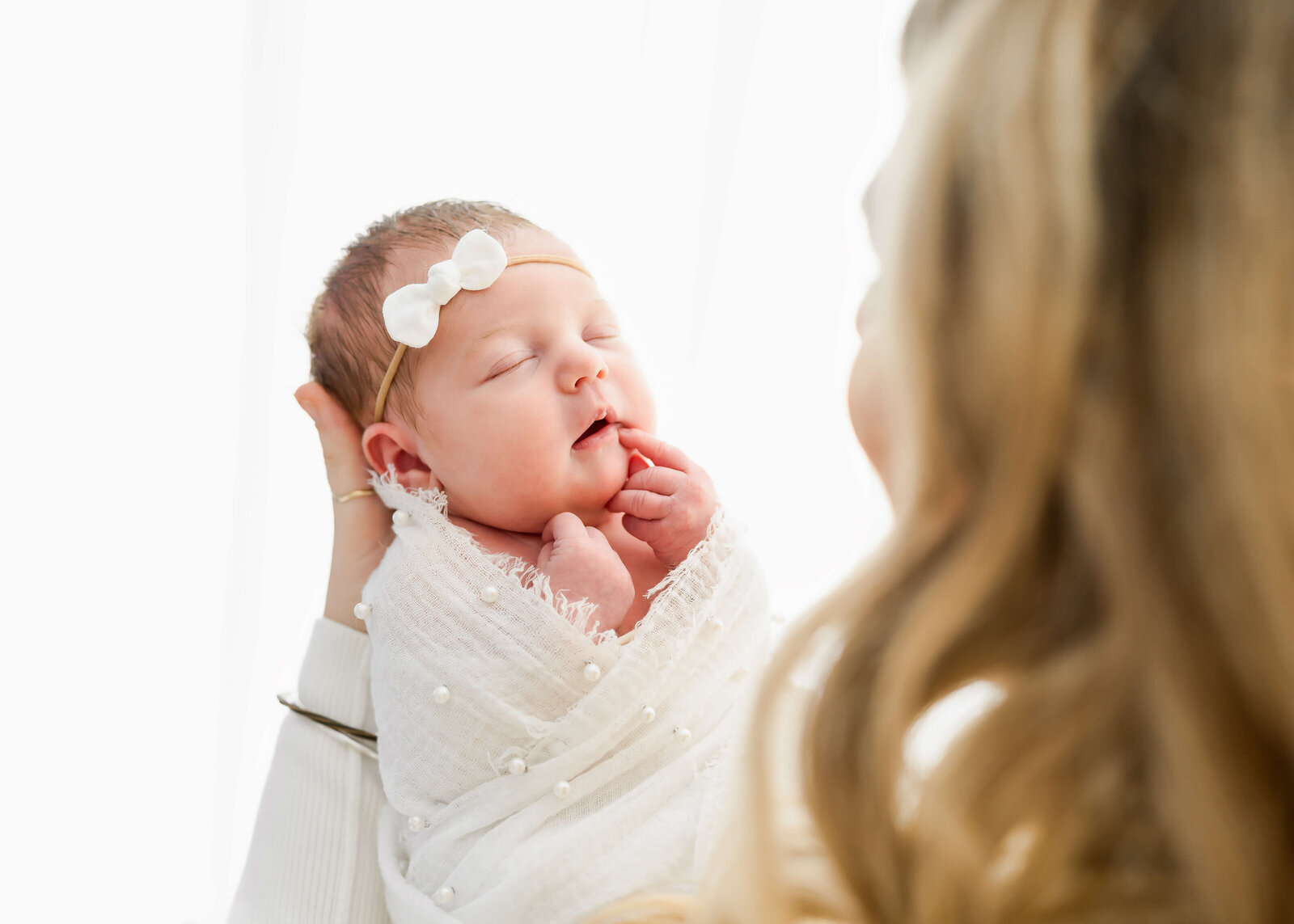 A newborn baby girl in a white swaddle sleeping in mom's hands with a white bow.