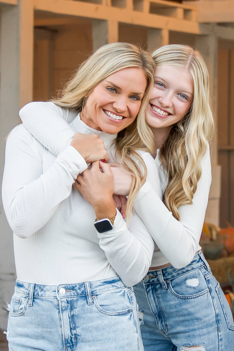 A teen girl hugs her mom from behind as they wear matching white tops and jeans.