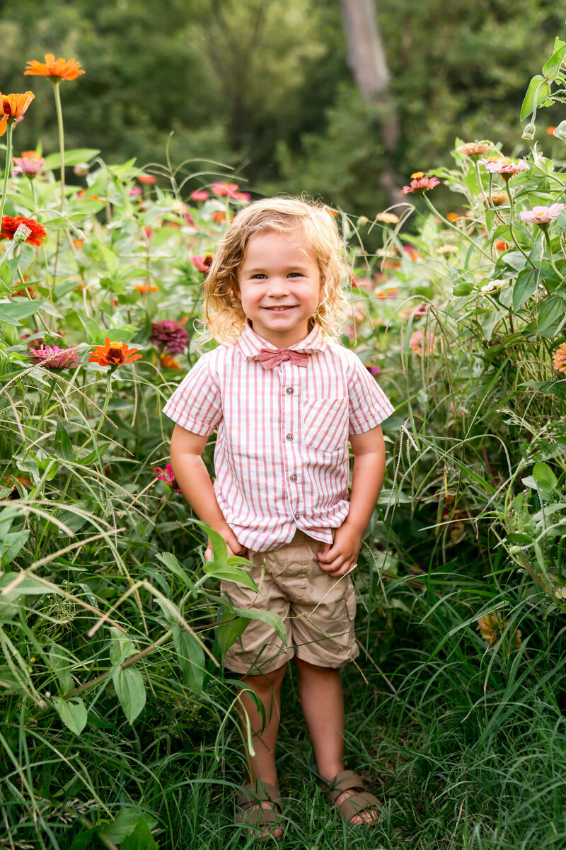 A toddler in a pink shirt and bowtie stands in a wildflower garden smiling.