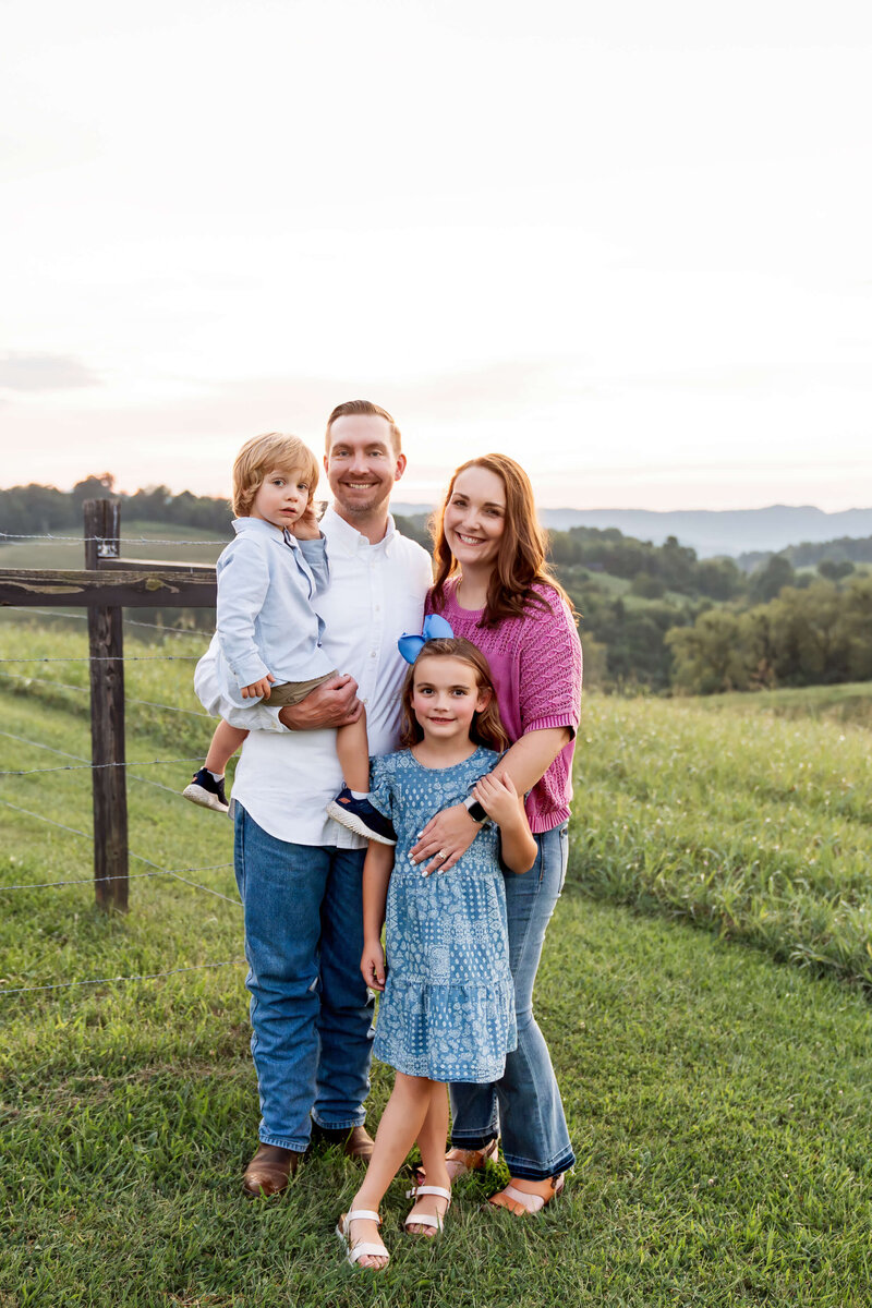 A mom and dad standing on a hilltop at sunset smiling with their toddler son and daughter.