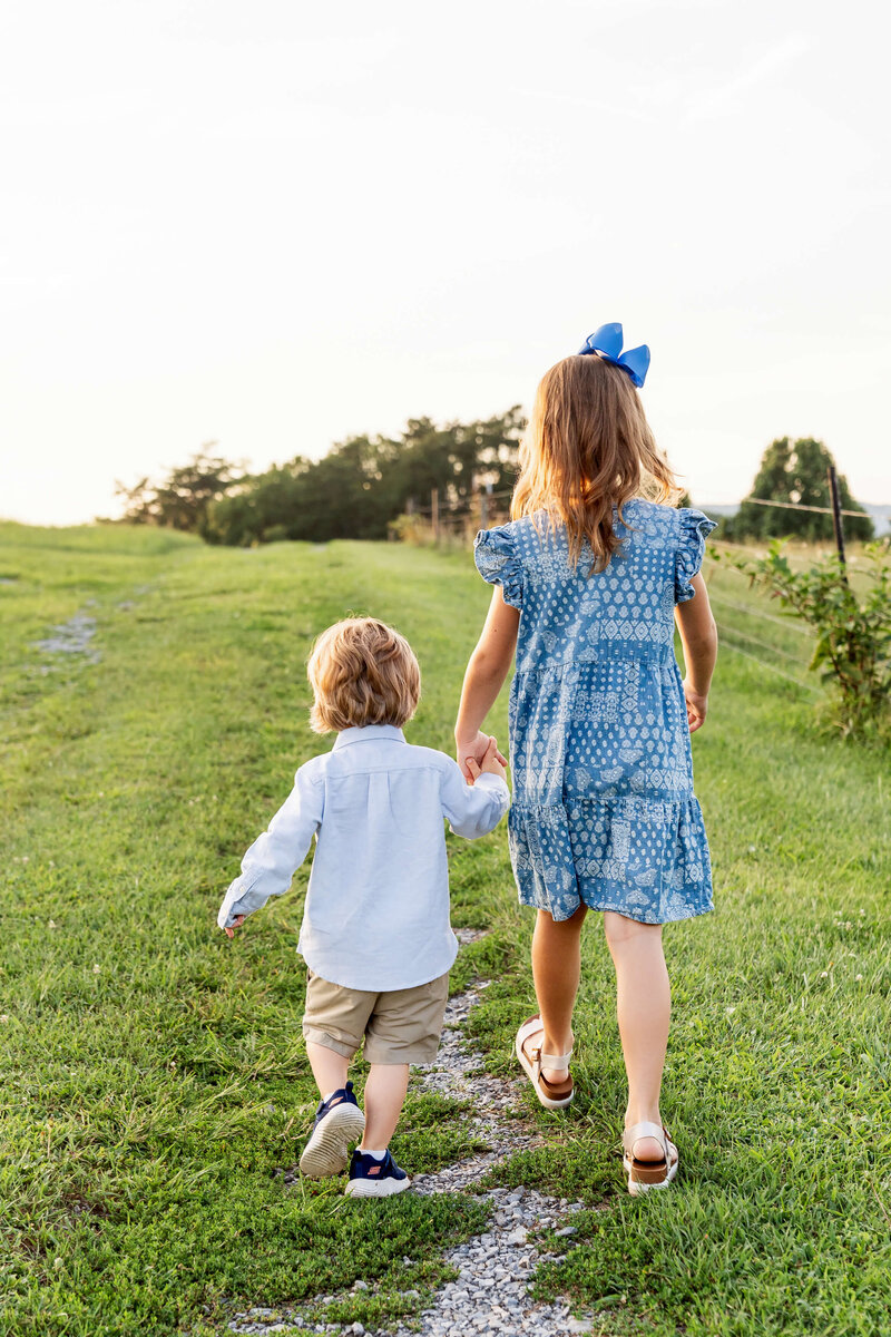 A toddler girl in a blue dress and bow walks up a hill trail holding hands with her little brother in white at sunset.