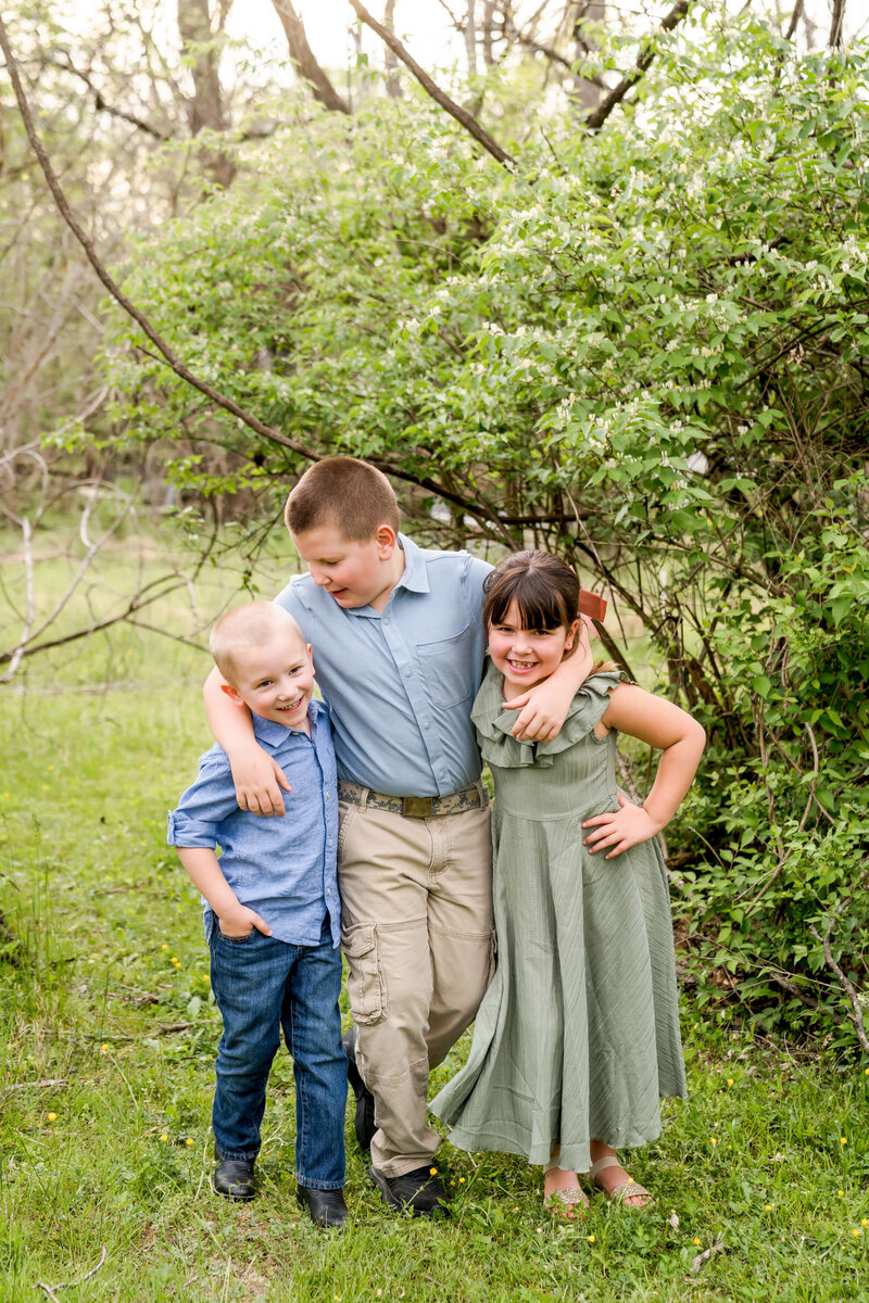 A young boy in a blue shirt hugs his two younger cousins in a park as guided by a Johnson City family photographer.