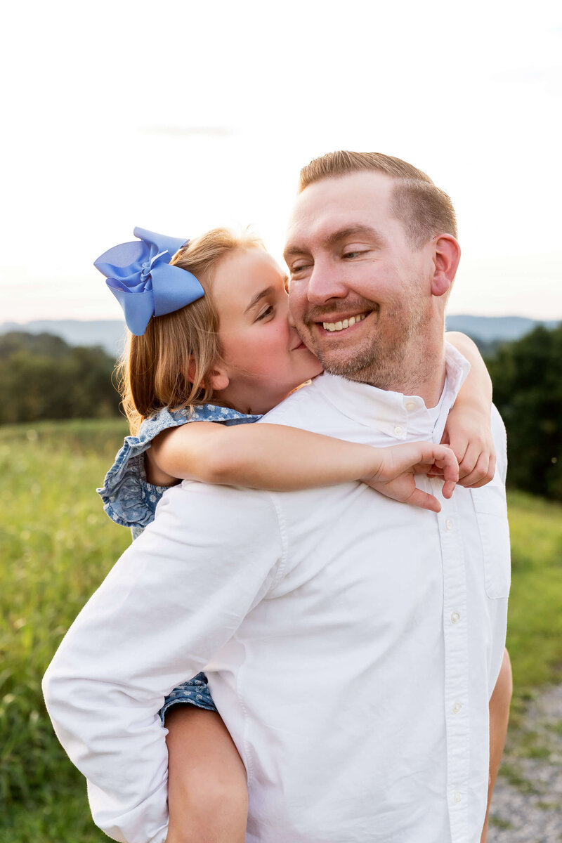 A toddler girl hangs on dad's back while kissing his cheek in a meadow from a Johnson City  family photography session.