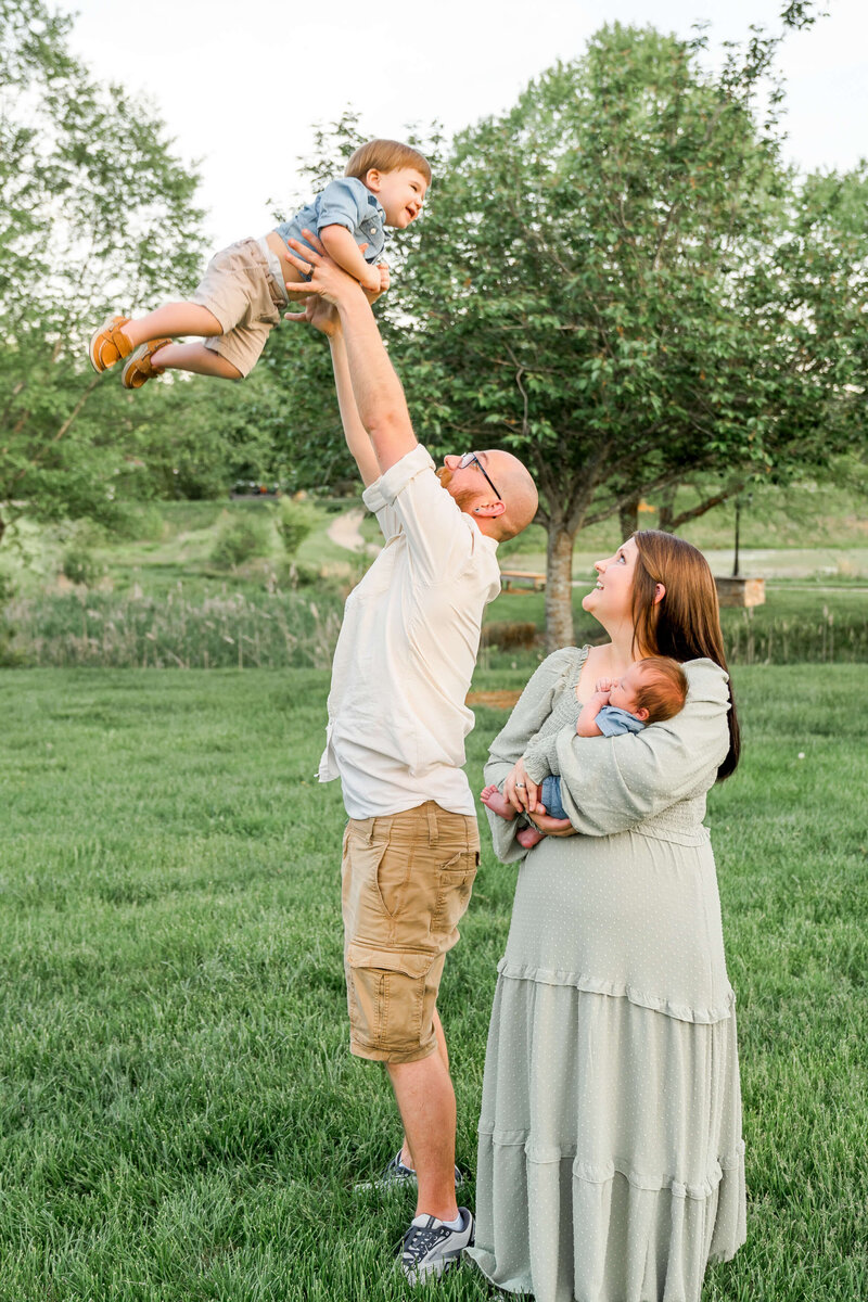A moment in a park lawn captured by a Johnson City family photographer of a dad in white tossing his toddler in the air as mom and infant watch.