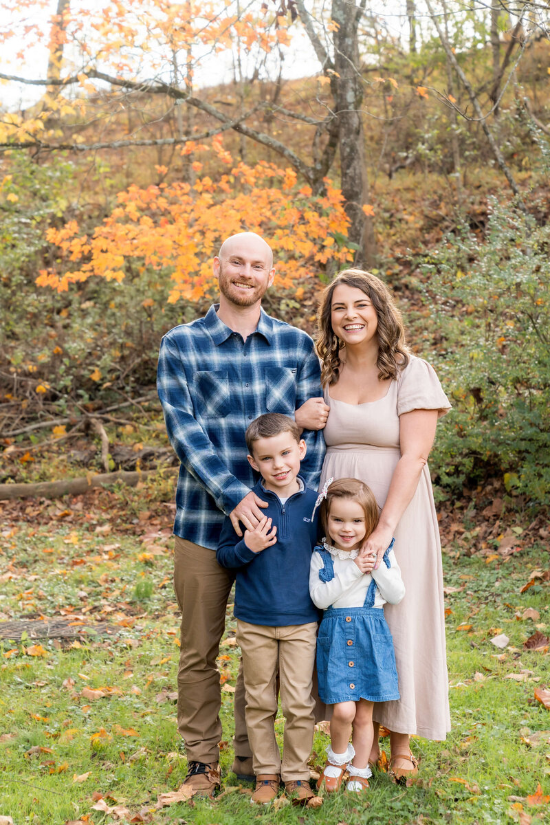 A mom and dad stand smiling in blue and cream with their toddler son and daughter in a lawn.