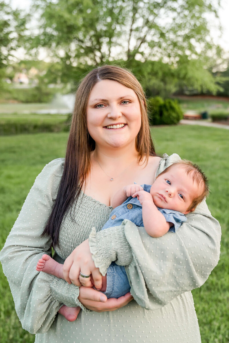 A happy mom in a green dress smiles while holding her infant in her arms in a park.