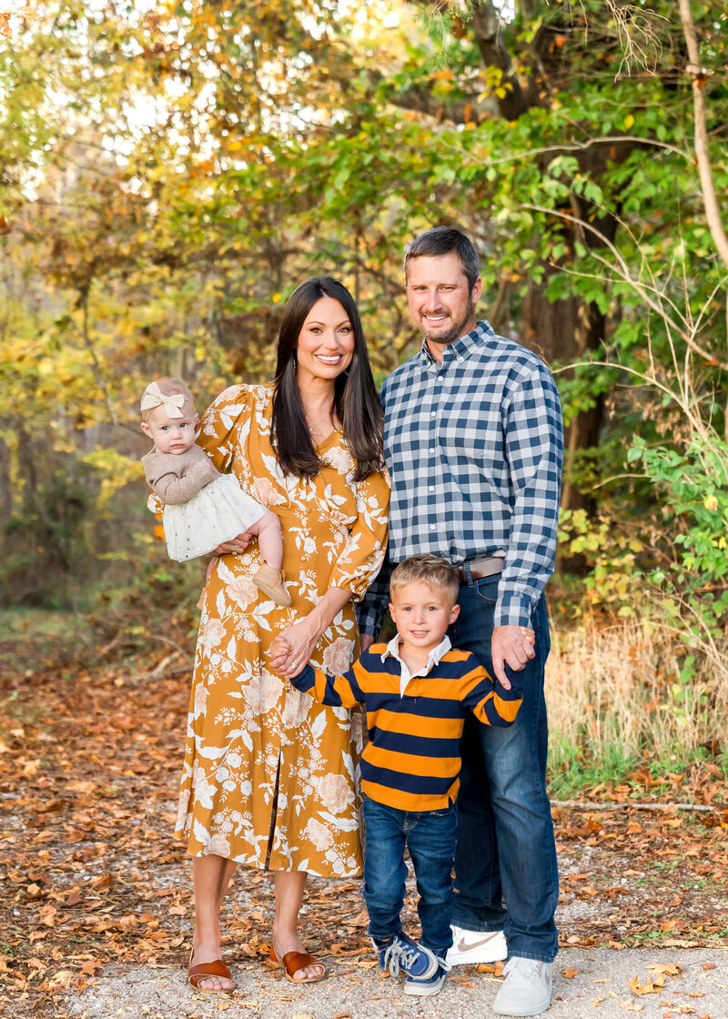 A toddler boy smiles while standing with mom and dad as baby sister sits in mom's arms all wearing blue and orange.