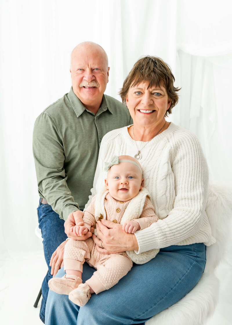 Happy grandparents smile while sitting with their baby granddaughter in her lap.