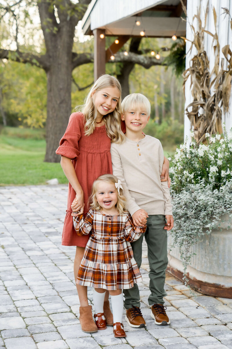 A young girl in a red dress hugs her smiling younger brother and sister on a patio by flowers.