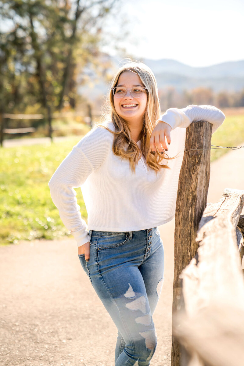 A teenager in a white blouse and jeans leans on a fence post smiling at sunset.