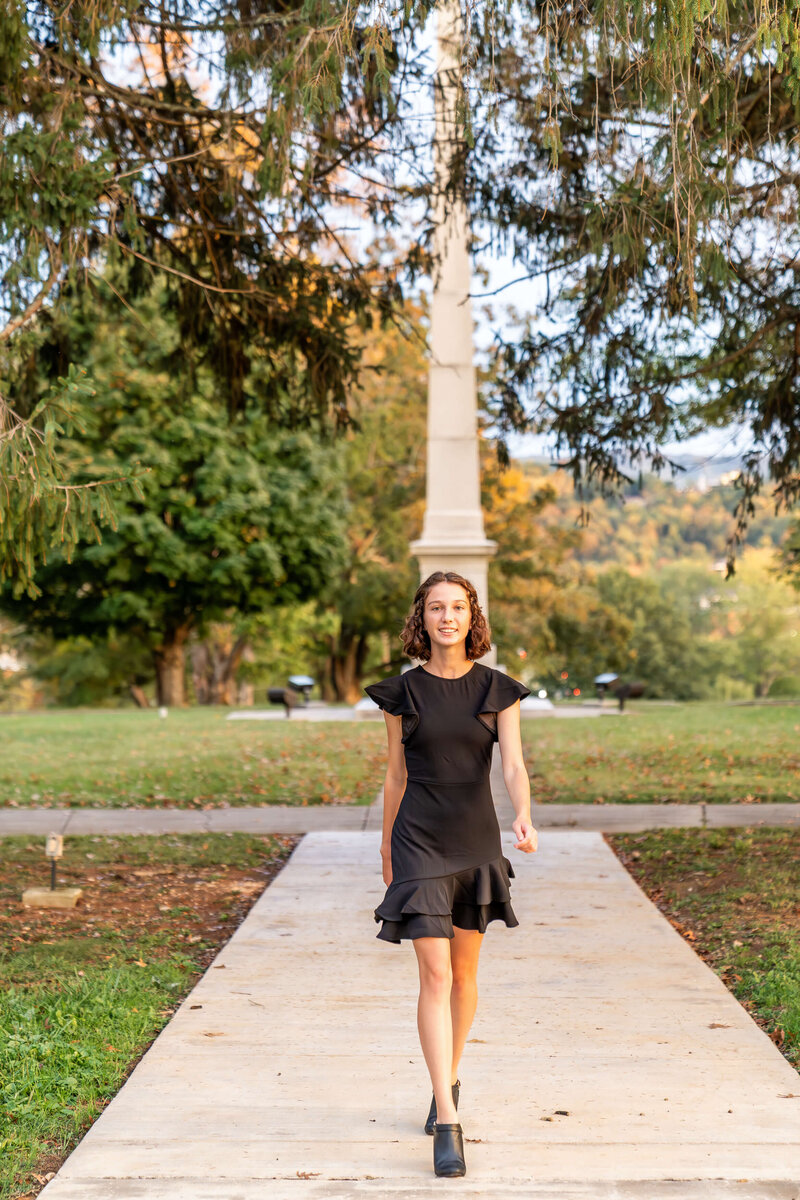 A high school senior walks in a park sidewalk by a monument at sunset in a black dress.
