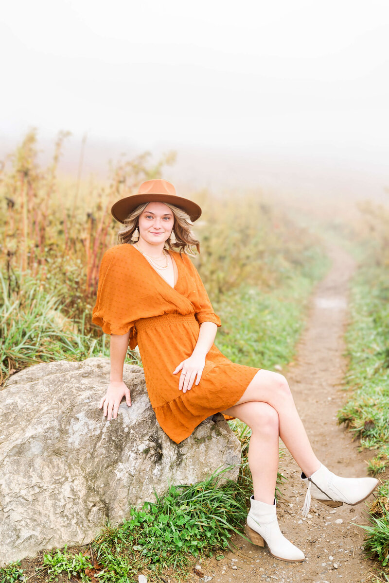 A high school grad in a rust colored dress sits on a boulder in a trail through tall golden grass.