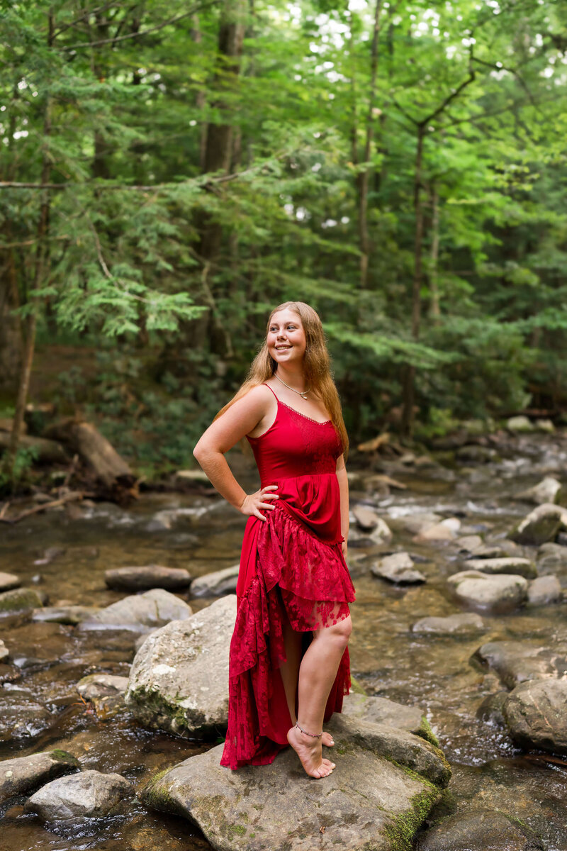 A high school senior in a red gown stands with a smile and hand on her hip in a creek.