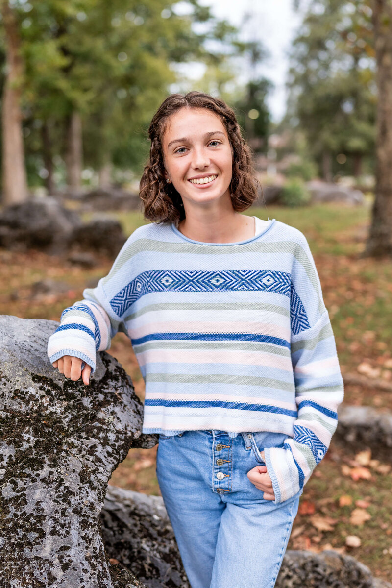 A woman smiles while leaning on a boulder in a blue and white sweater with a thumb in her pocket during her Johnson City senior grad photography session.