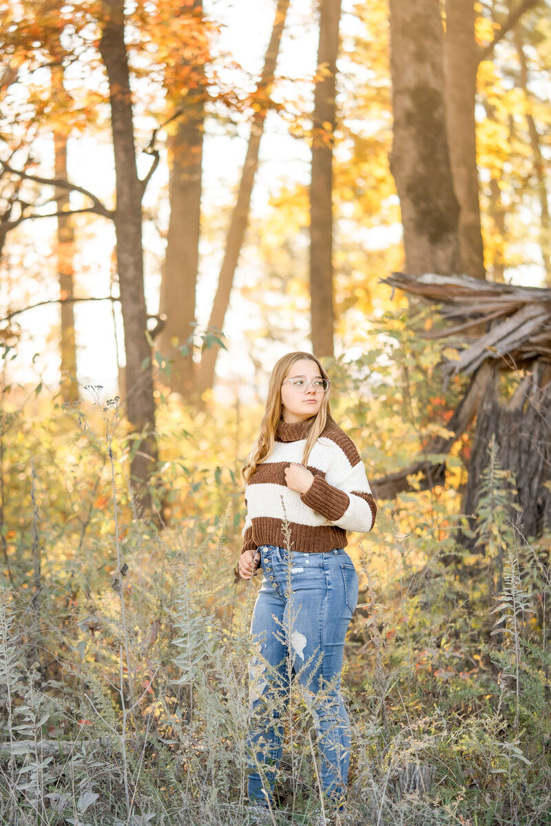 A teenager in a brown and white blouse hikes in a forest at sunset while looking over her shoulder.