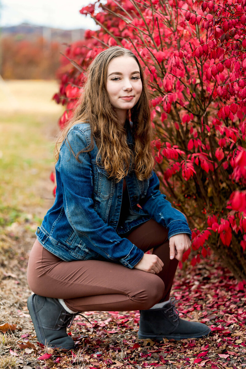 A high school senior in a denim jacket and brown pants kneels next to the red flowers with a big smile.