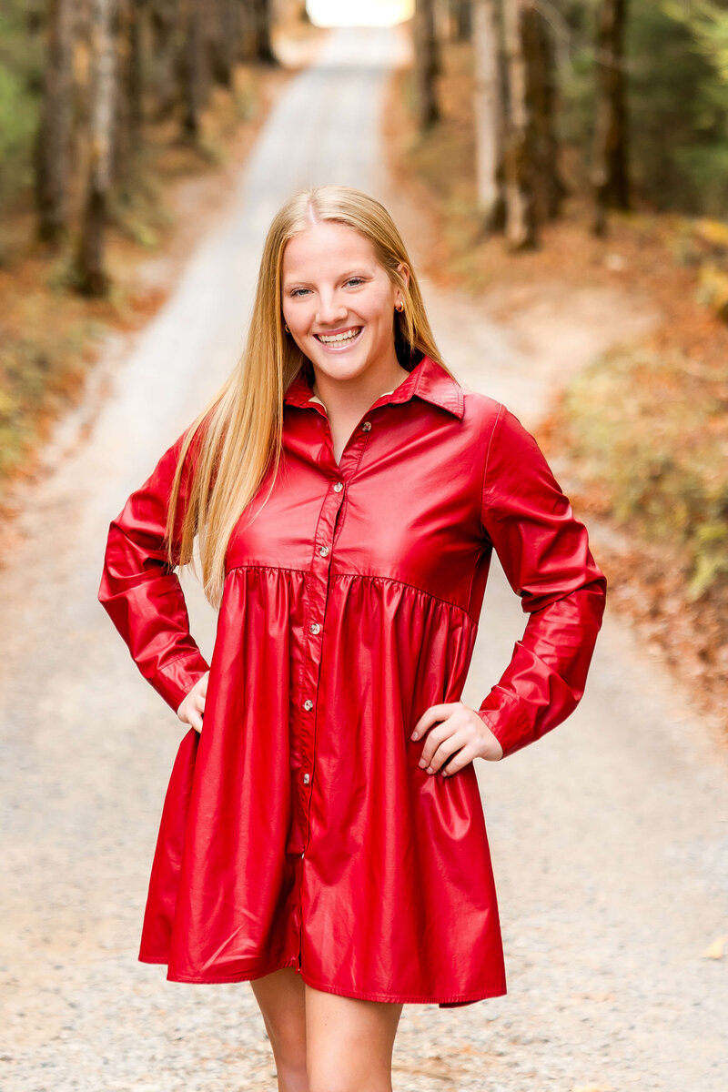 A grad smiles big while standing in a park trail in a red dress.