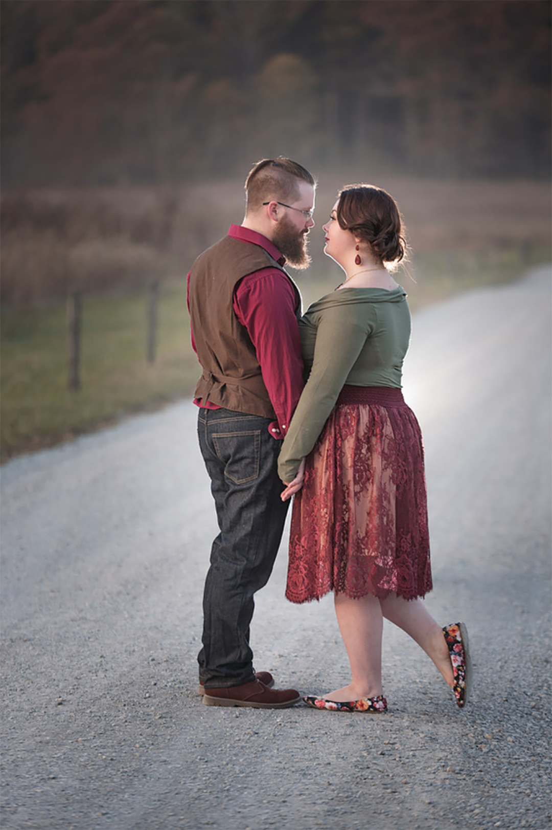 An expecting couple in green and brown snuggle holding hands under the bump in a park in the fall captured by their Johnson City maternity photographer.