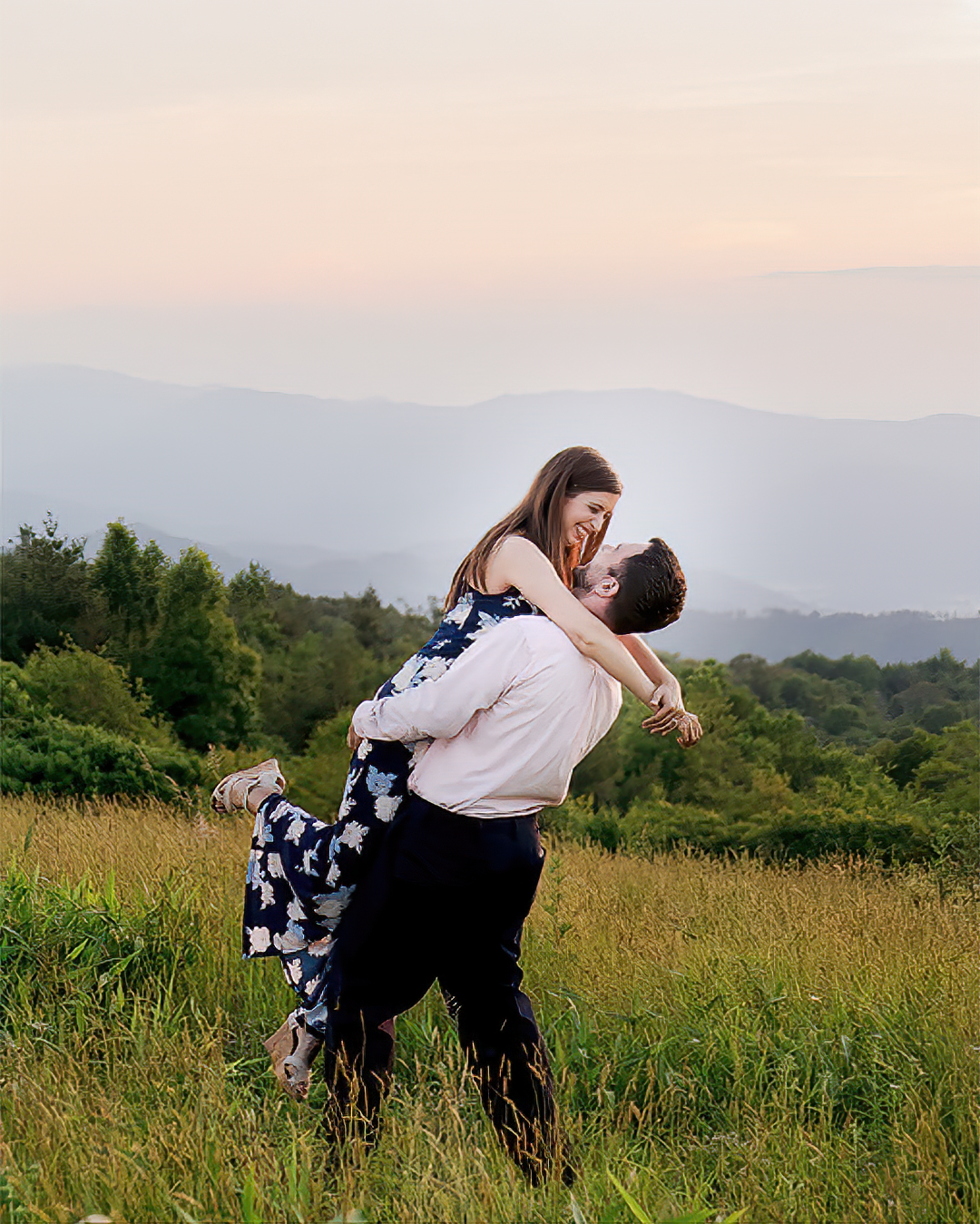 A man lifts a woman in his arms in a grassy meadow during an engagement session near Johnson City.