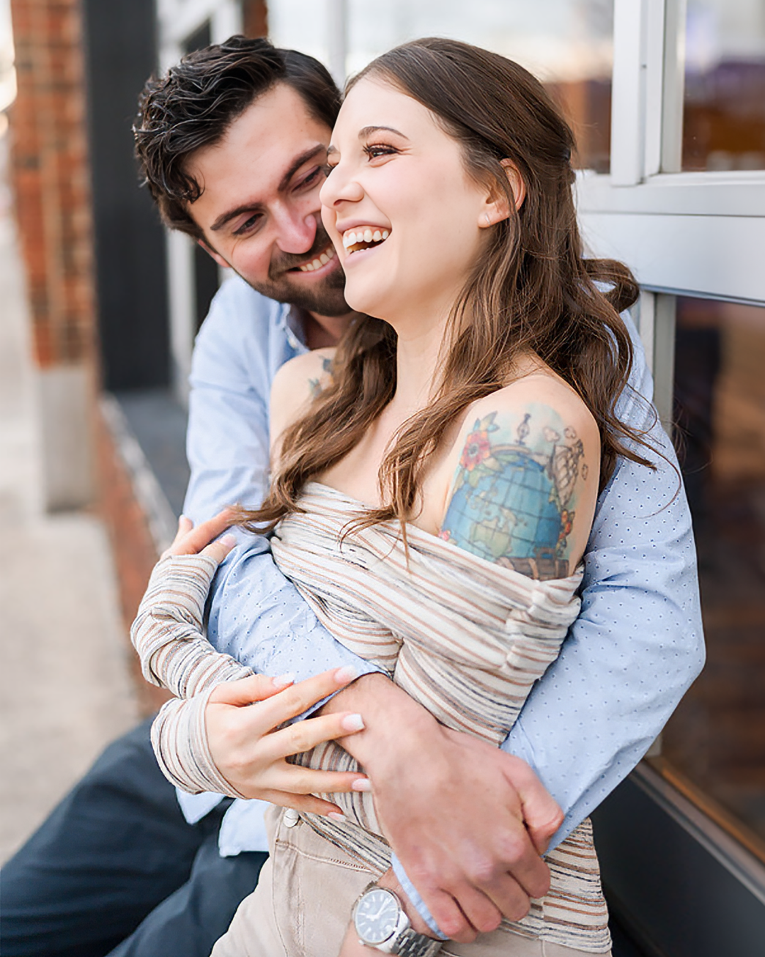 A couple cuddles together against a brick building during their engagement session.
