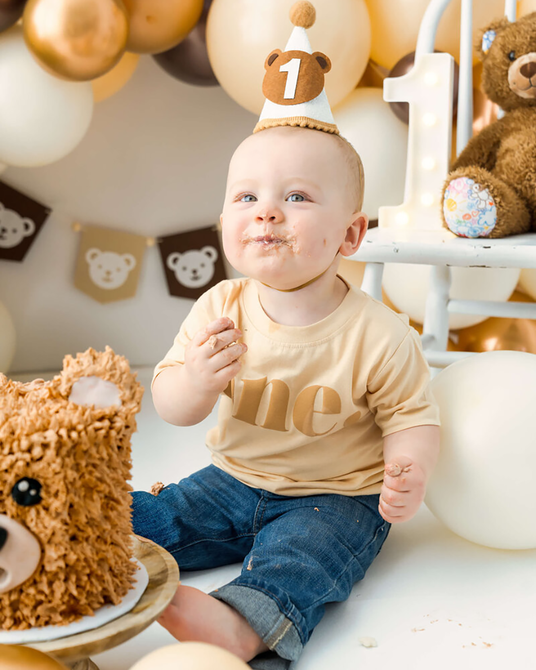 A one year old baby boy with a birthday hat and a teddy bear cake eats and smiles with birthday balloons at a milestone session.