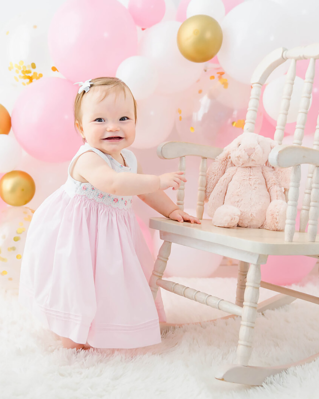 A little girl dressed in pink stands up holding onto a white rocking chair with a bunny sitting on it and pink and white balloons in the background at a photo session in Johnson City.