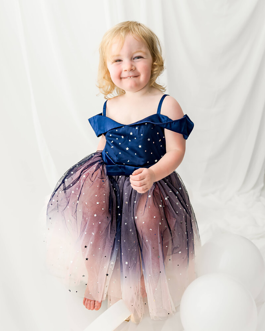 A toddler girl in a blue and pink sparkling dress smiles in a Johnson City newborn photographer's studio.