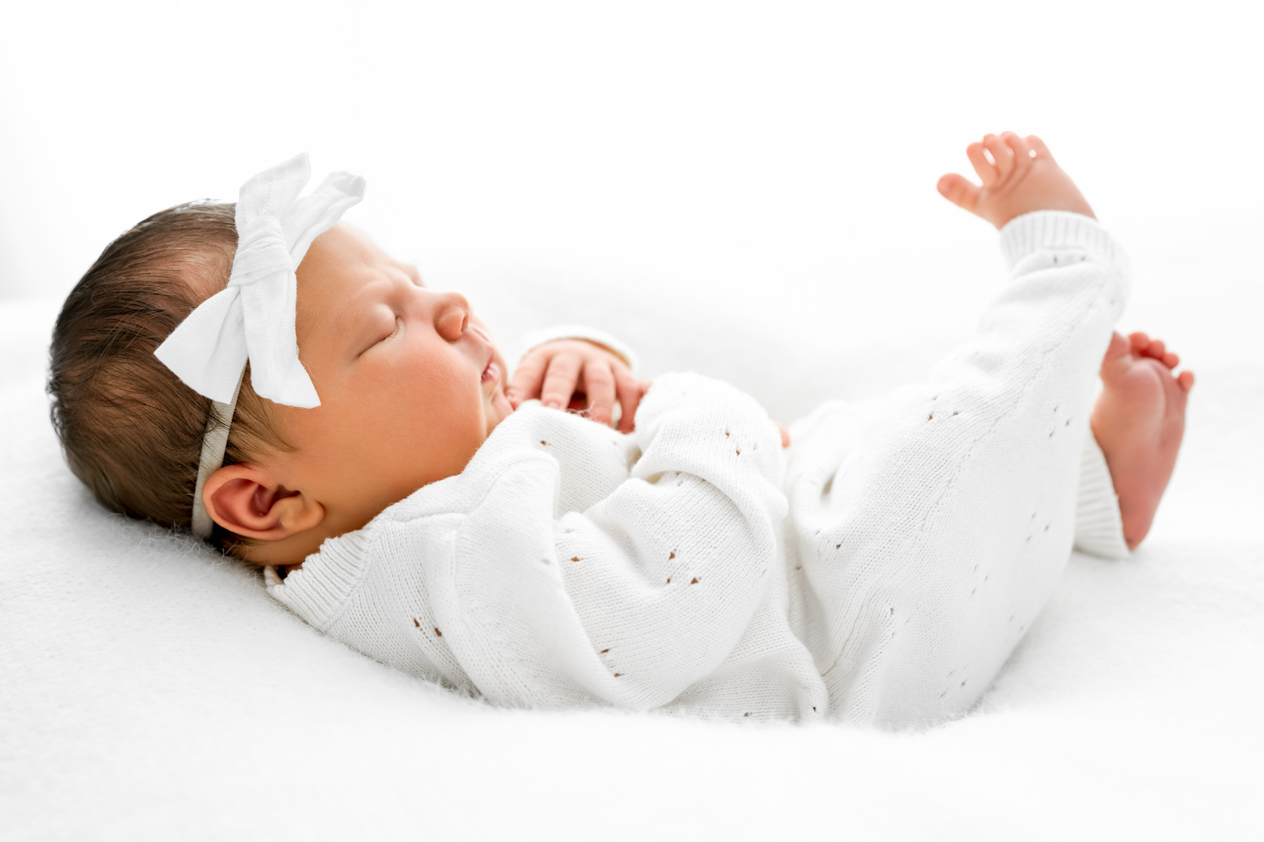 Sleeping newborn baby girl wearing a white bow and white knit outfit on a soft white blanket.
