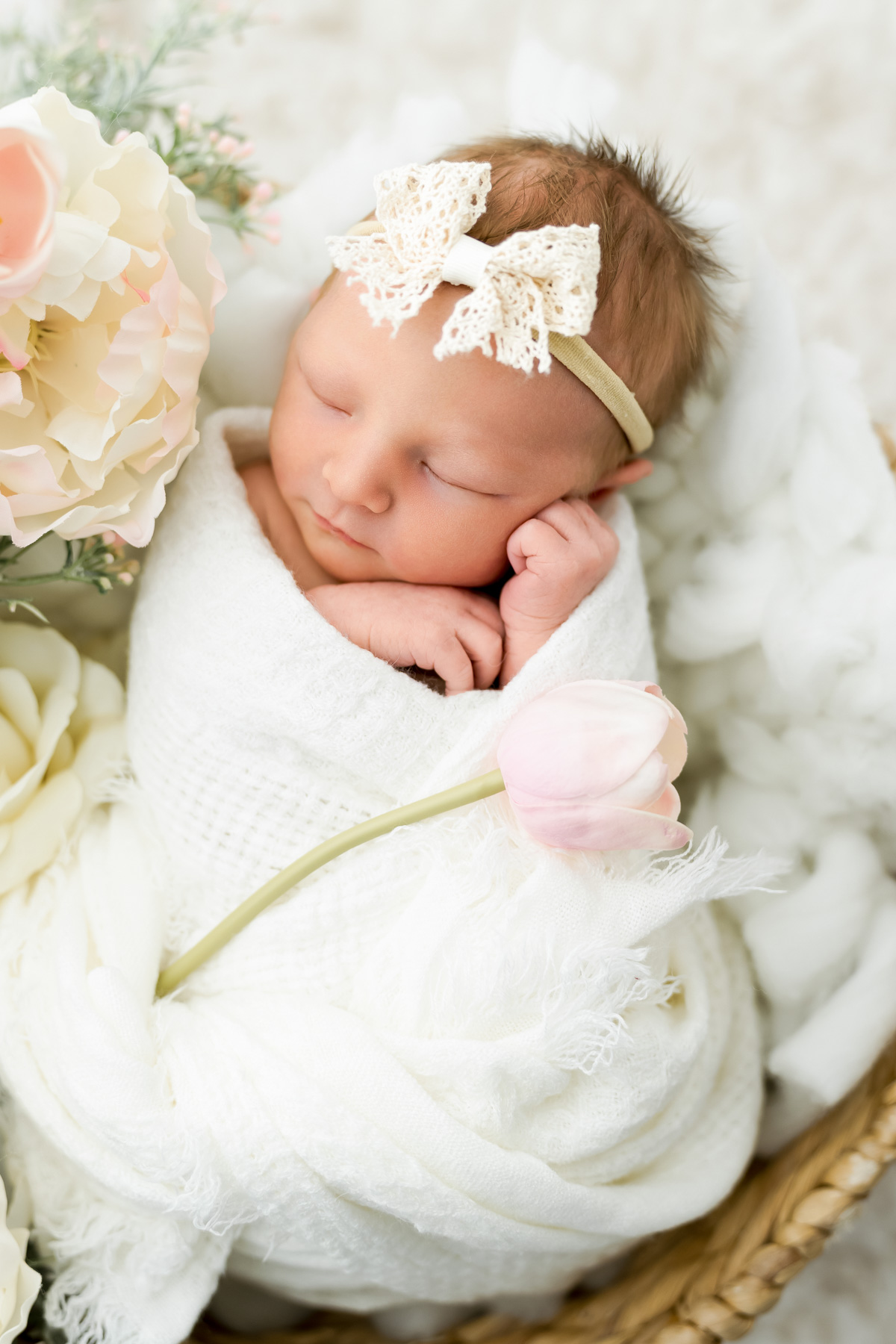 Sleeping newborn baby girl wrapped in white with a lace bow, posed beside soft pink flowers.