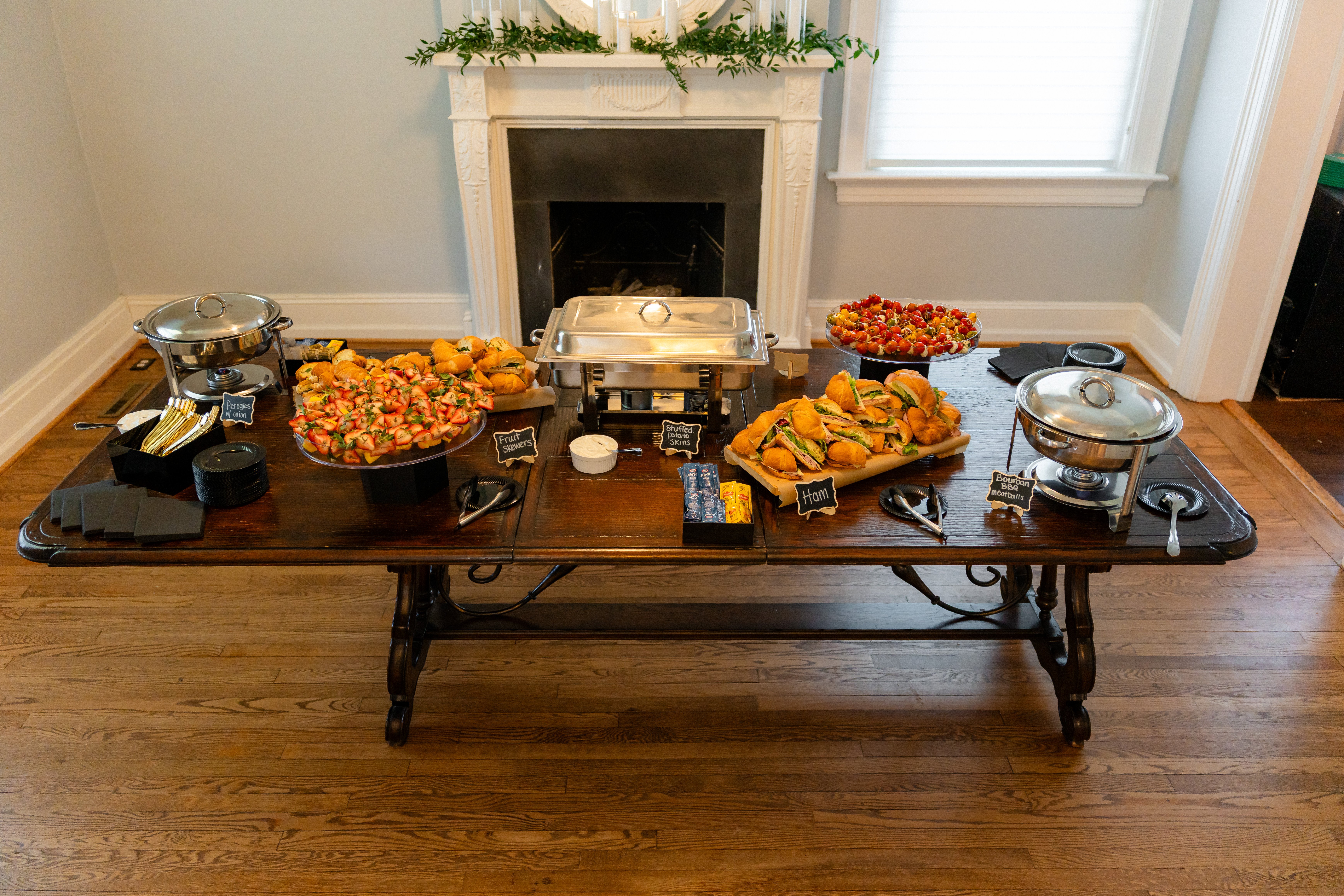 Full catering buffet displayed on an antique wood table in front of a decorated fireplace at Bent Oaks Manor